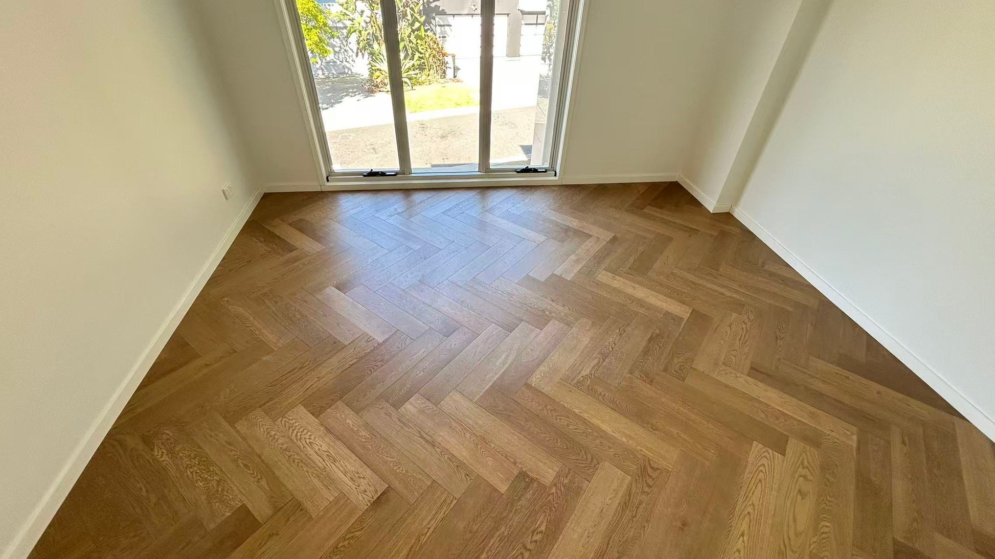 Coral Sand iDesign Herringbone Flooring 15/3mm installed in an empty sunlit room with large sliding glass doors and white walls.