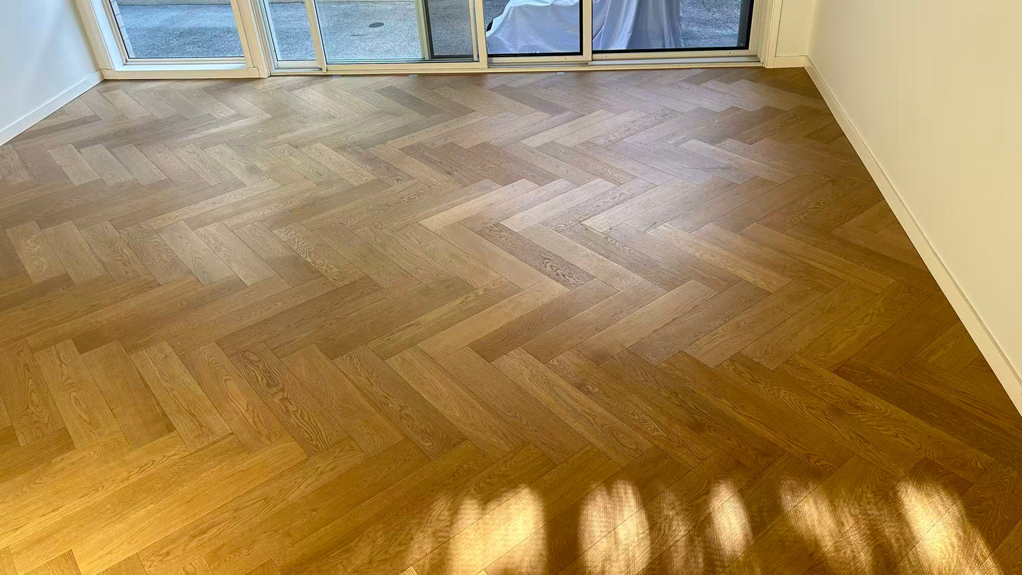 Coral Sand iDesign Herringbone Flooring 15/3mm installed in a sunlit empty room with sliding glass doors, showcasing a warm golden-brown oak herringbone pattern.