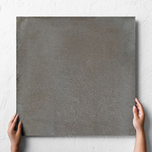 Hand holding a single Cotto Concrete Matt 600x600mm tile against a white rendered wall, showing the matt surface