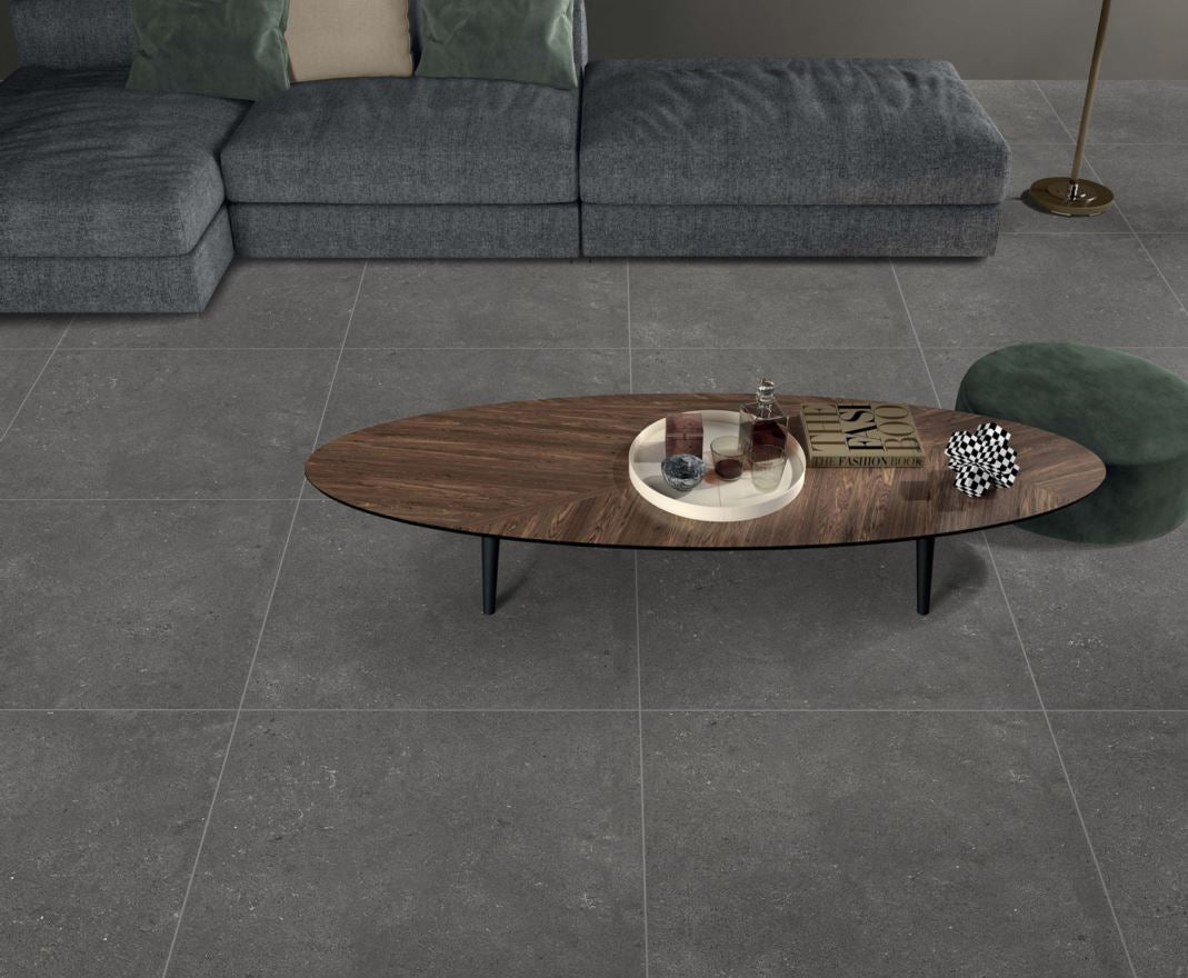 Esmal Charcoal 600x600mm porcelain floor tiles installed in a modern living room with a walnut oval coffee table and grey sectional sofa.