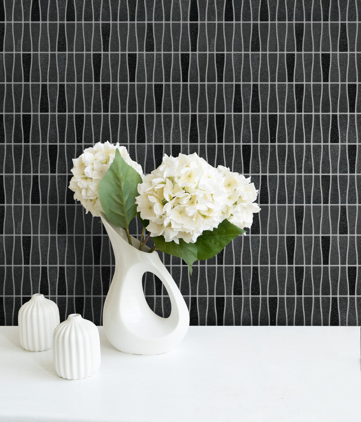 Forever Black Recycled Glass Shading Wedge 290x309mm tile wall styled with a sculptural white vase holding white hydrangeas and small ribbed ceramic vessels on a white surface.