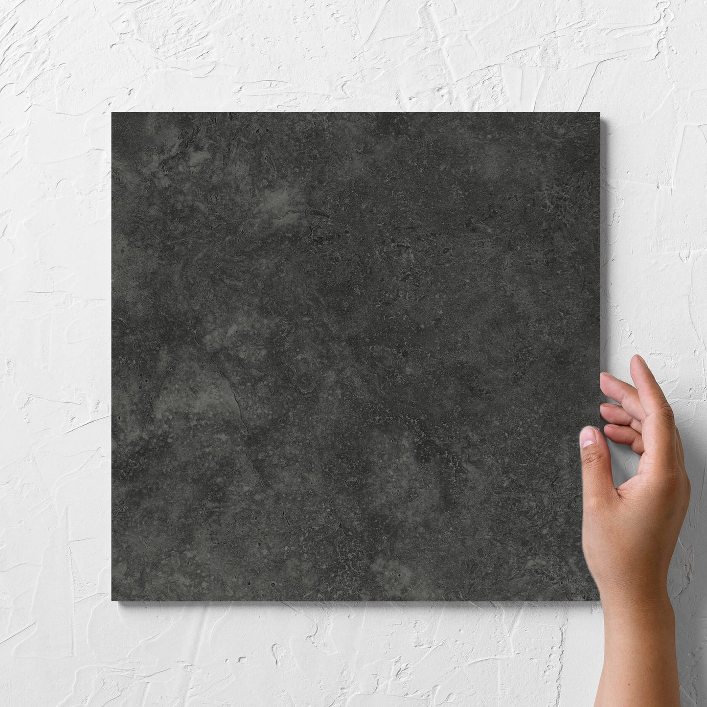 Hand holding a single Fossil Stone Coal Matt 300x300mm tile against a white rendered wall, showing the matt surface