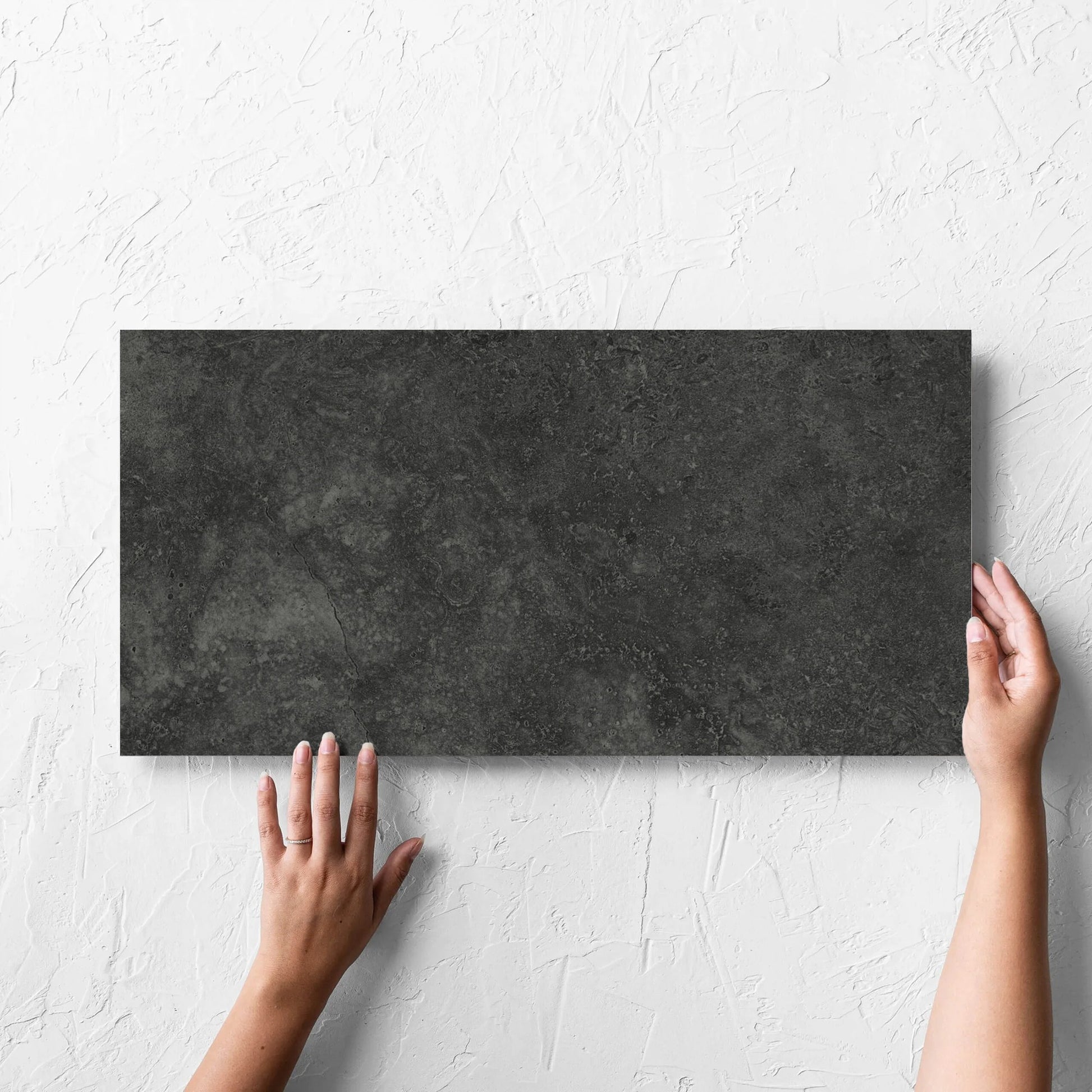 Hand holding a single Fossil Stone Coal Matt 300x600mm tile against a white rendered wall, showing the matt surface