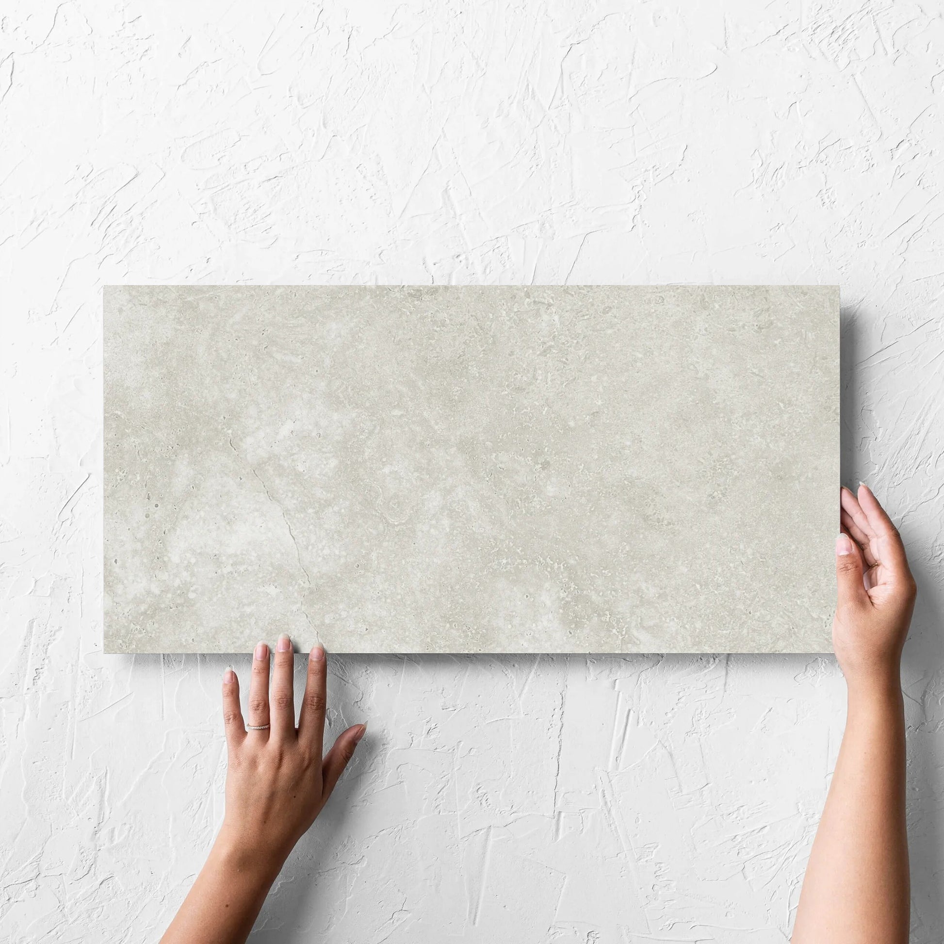 Hand holding a single Fossil Stone Sand Matt 300x600mm tile against a white rendered wall, showing the matt surface
