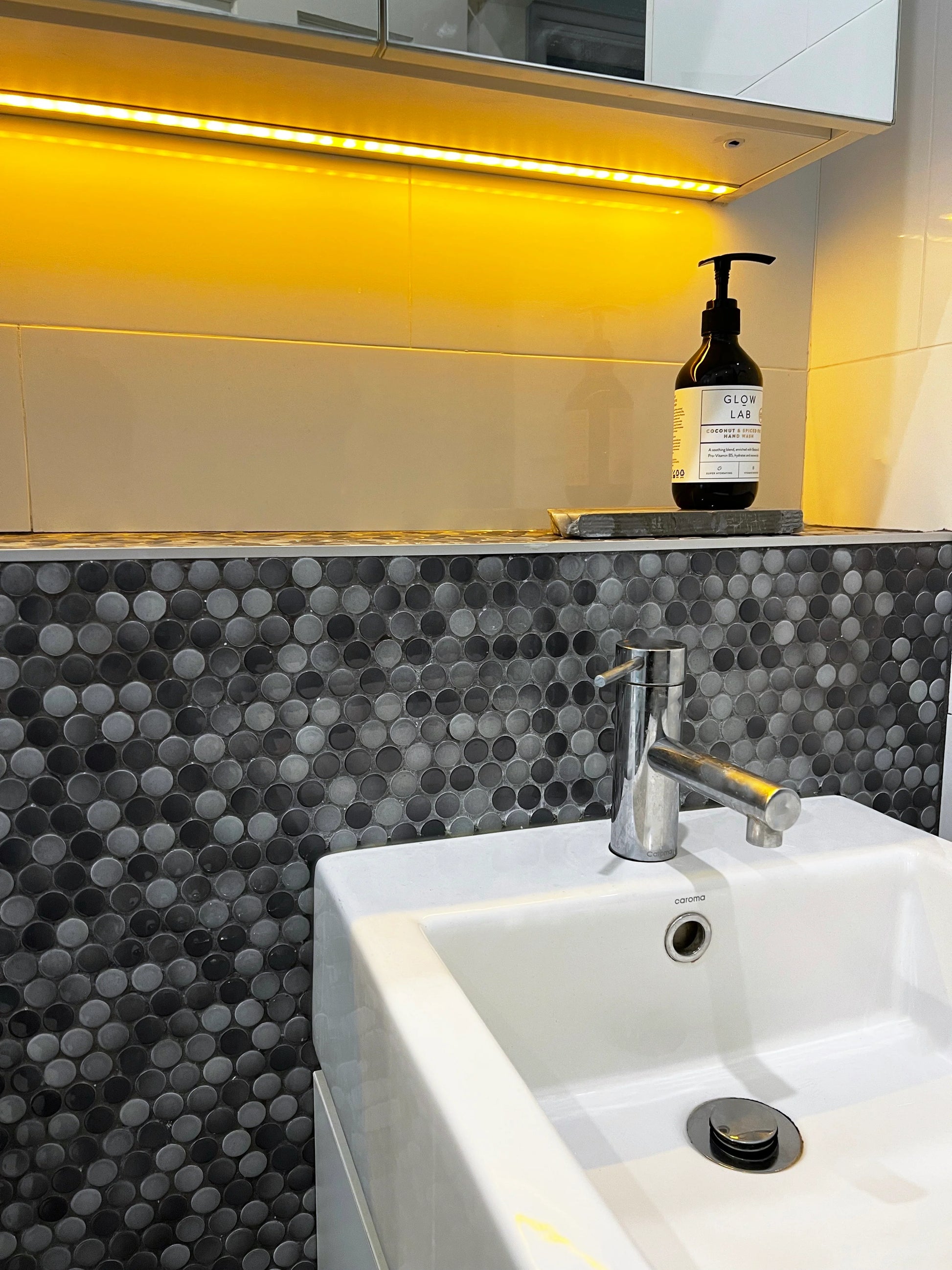 Grey Gloss Diameter Glazed Porcelain Penny Round 19mm tiles installed as a bathroom vanity splashback, paired with a white Caroma basin, chrome mixer tap, and warm under-cabinet LED lighting.
