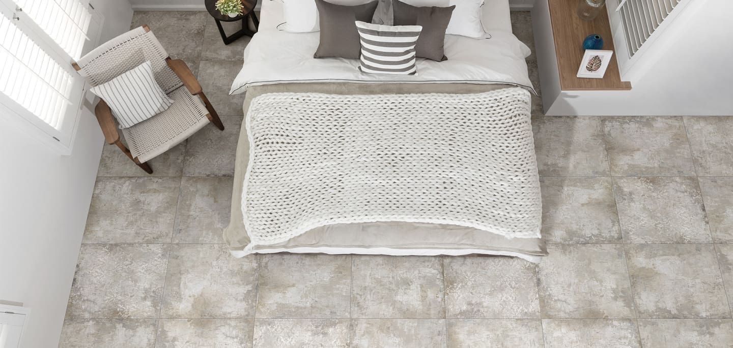 Grunge Beige Rectified 600x600mm tiles covering a bright bedroom floor, viewed from above with a styled bed and woven accent chair.