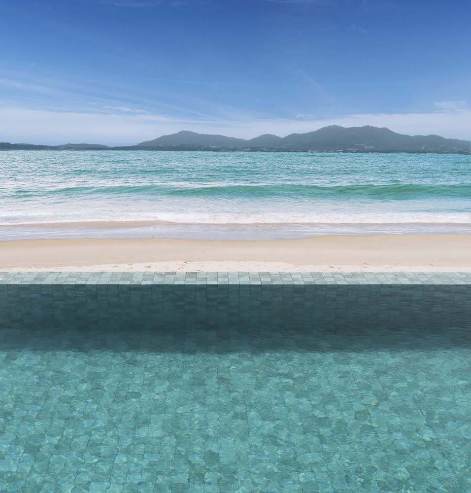 Hammond Green Mosaic 97x97mm tiles lining an infinity pool edge with a turquoise ocean, sandy beach, and mountain backdrop beyond.