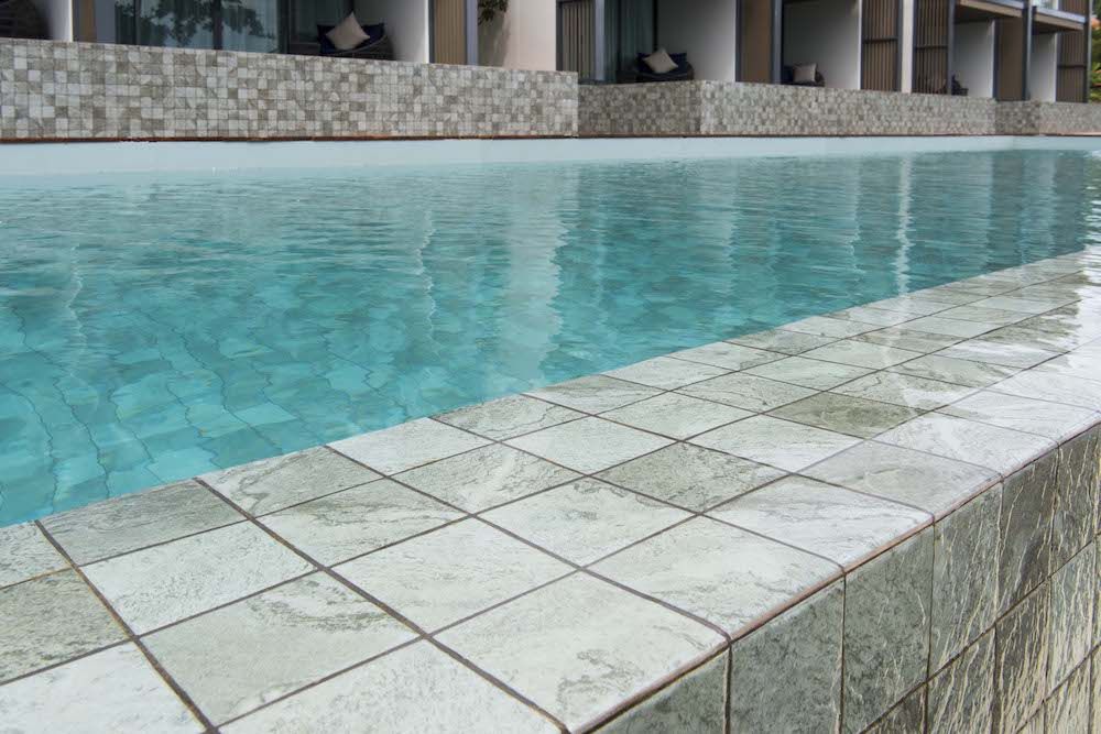 Hammond Green Mosaic 97x97mm tiles lining the coping edge of a luxury outdoor infinity pool with clear turquoise water and resort building backdrop.
