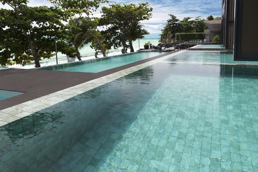 Hammond Green Mosaic 97x97mm tiles lining a luxury resort infinity pool with turquoise water, tropical trees, and ocean views beyond.