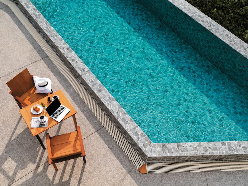 Hammond Grey Mosaic 97x97mm tiles lining a luxury swimming pool edge beside a timber outdoor table with laptop and croissant.