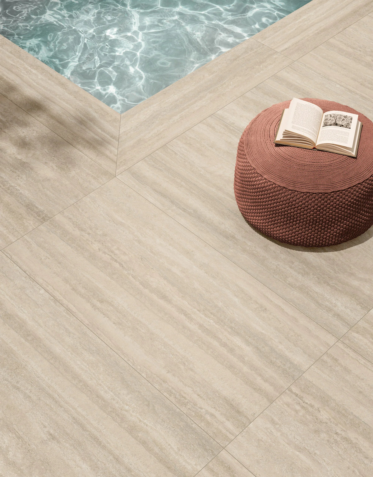 Ic.Verso Classico Gr 600x1200mm large-format beige stone-look tiles installed on a poolside patio with a woven terracotta ottoman and open book.