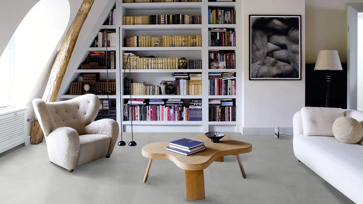 Imperial Light Grey Matt 600x600mm tiles laid as a living room floor beneath a boucle armchair, organic cork coffee table, and floor-to-ceiling white bookcase.