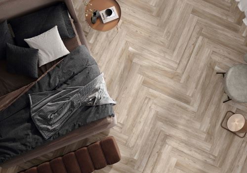 inout Olson Almond 230x1200mm porcelain tiles laid in a herringbone pattern on a bedroom floor beside a dark upholstered bed and timber side table.