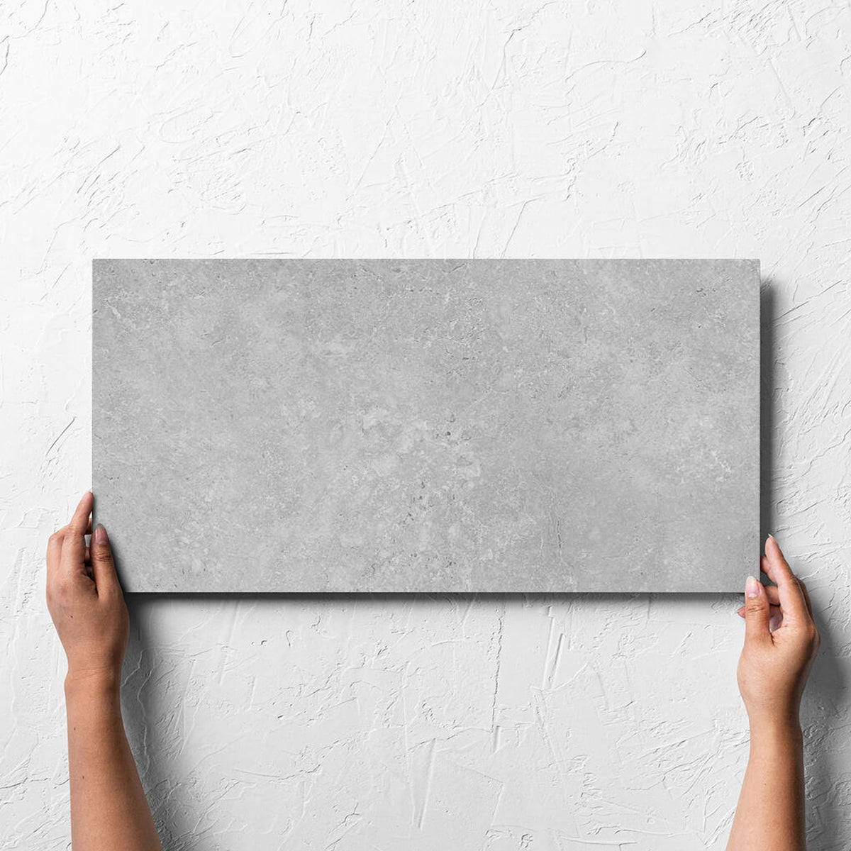 Hand holding a single Kross Light Grey Matt Smooth Grip 300x600mm tile against a white rendered wall, showing the tile surface