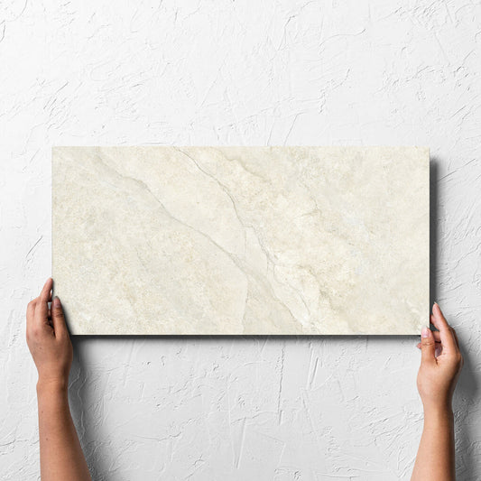 Hand holding a single Lakestone Sand Matt 300x600mm tile against a white rendered wall, showing the matt surface