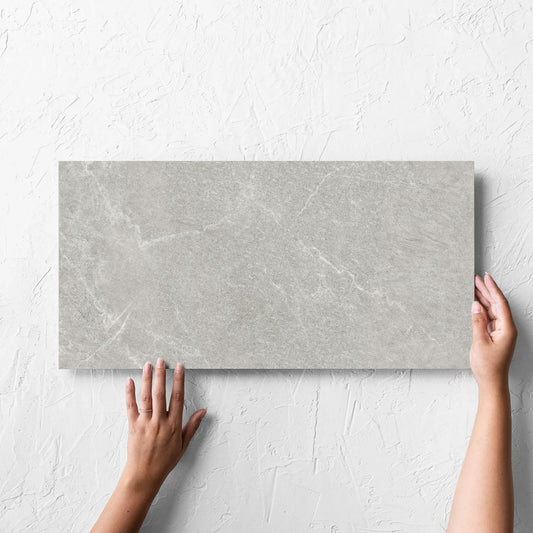 Hand holding a single Lava Ash Amber 300x600mm tile against a white rendered wall, showing the ash matt surface