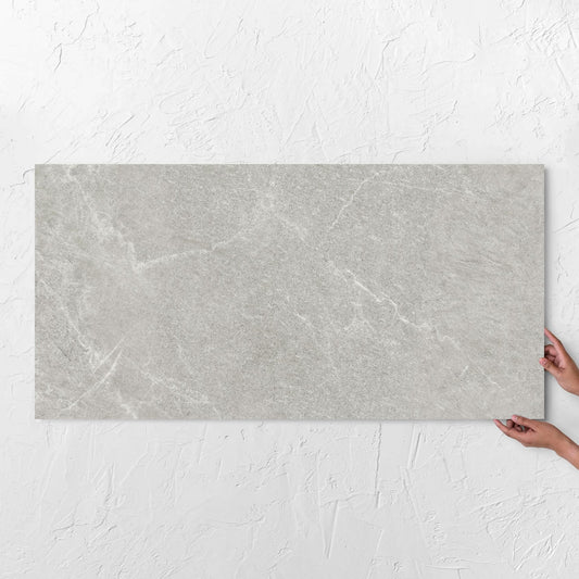 Hands holding a Lava Ash Amber 600x1200mm tile against a white rendered wall, showing the ash surface
