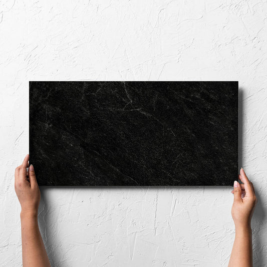 Hand holding a single Lava Black Amber 300x600mm tile against a white rendered wall, showing the lappato surface