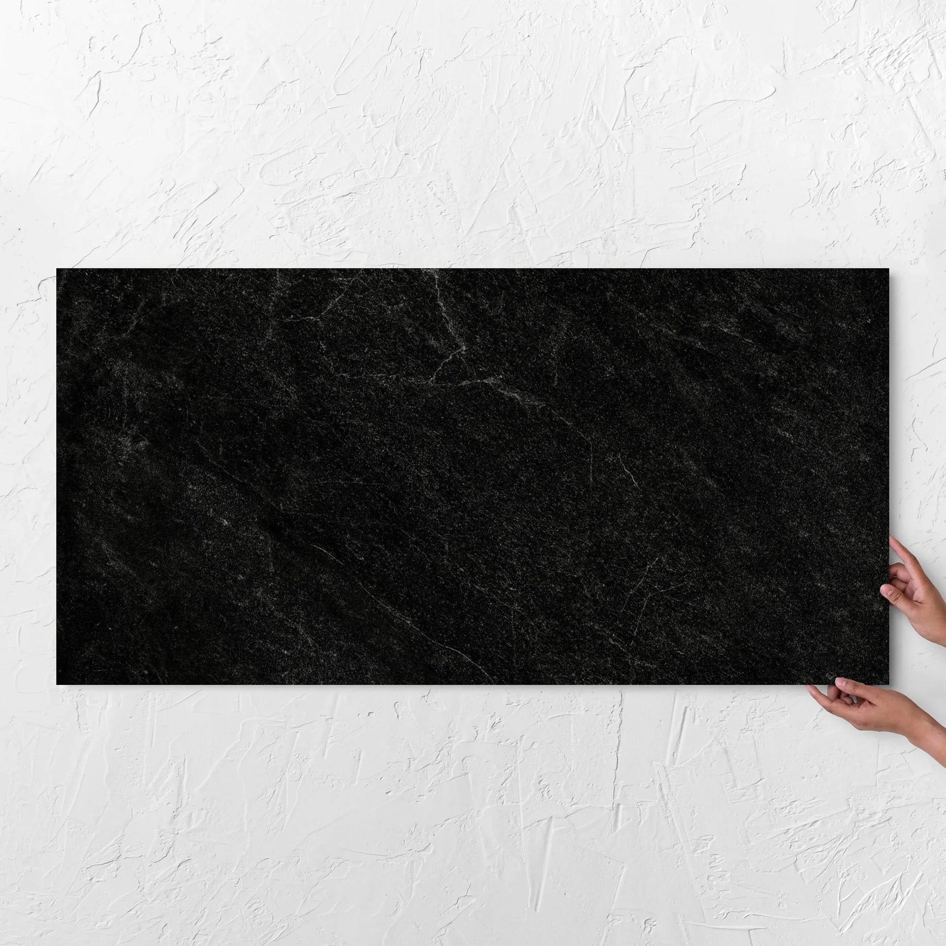 Hands holding a Lava Black Amber 600x1200mm tile against a white rendered wall, showing the black surface