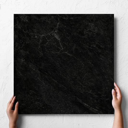 Hand holding a single Lava Black Rect Inout 600x600mm tile against a white rendered wall, showing the tile surface