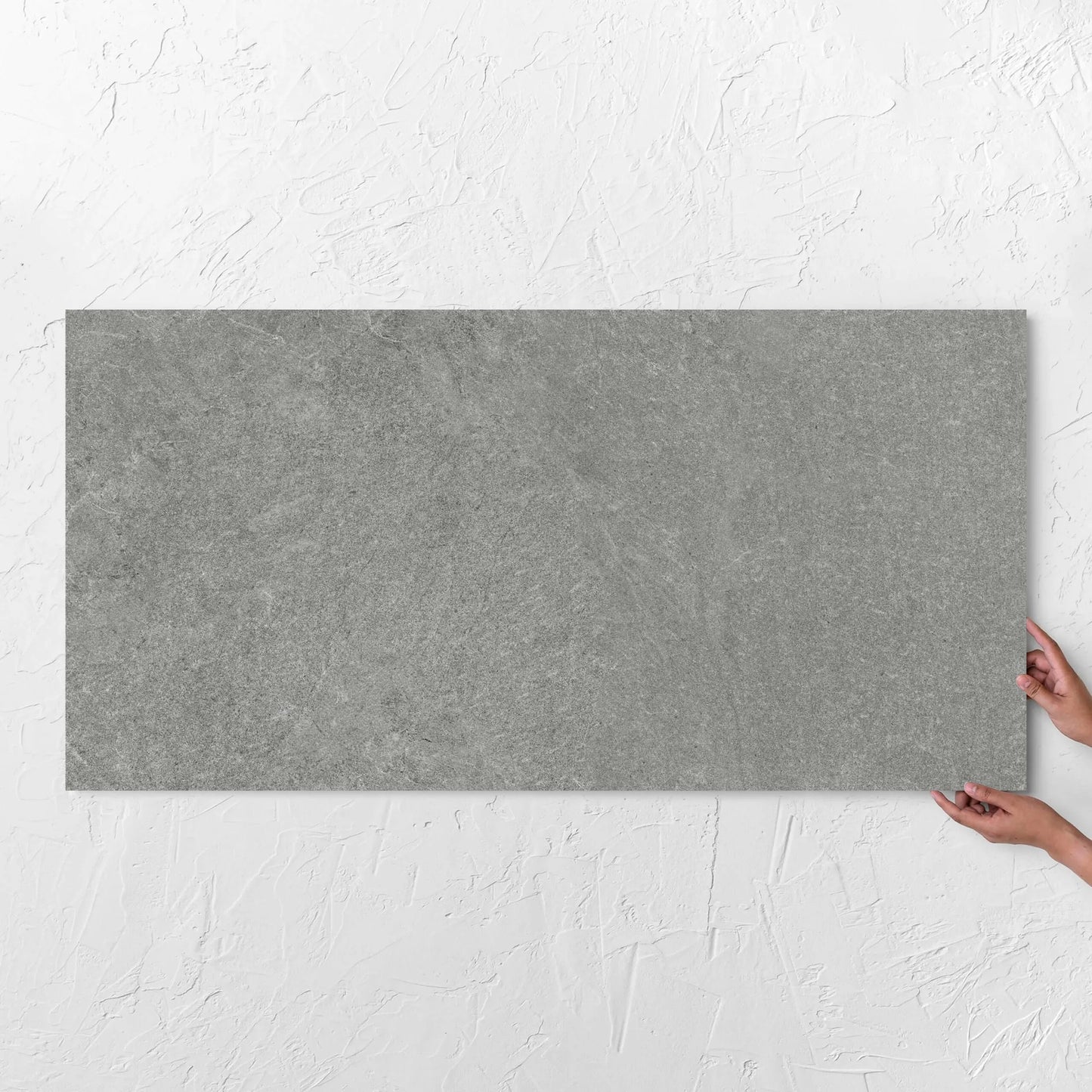 Hands holding a Lava Grey Amber 600x1200mm tile against a white rendered wall, showing the grey surface