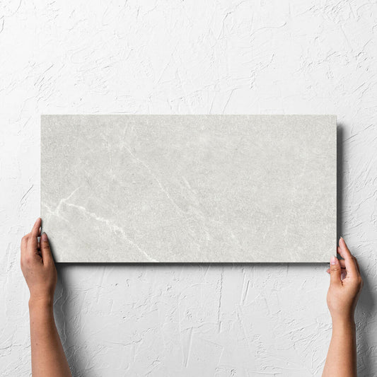 Hand holding a single Lava Warm Grey Amber 300x600mm tile against a white rendered wall, showing the lappato surface