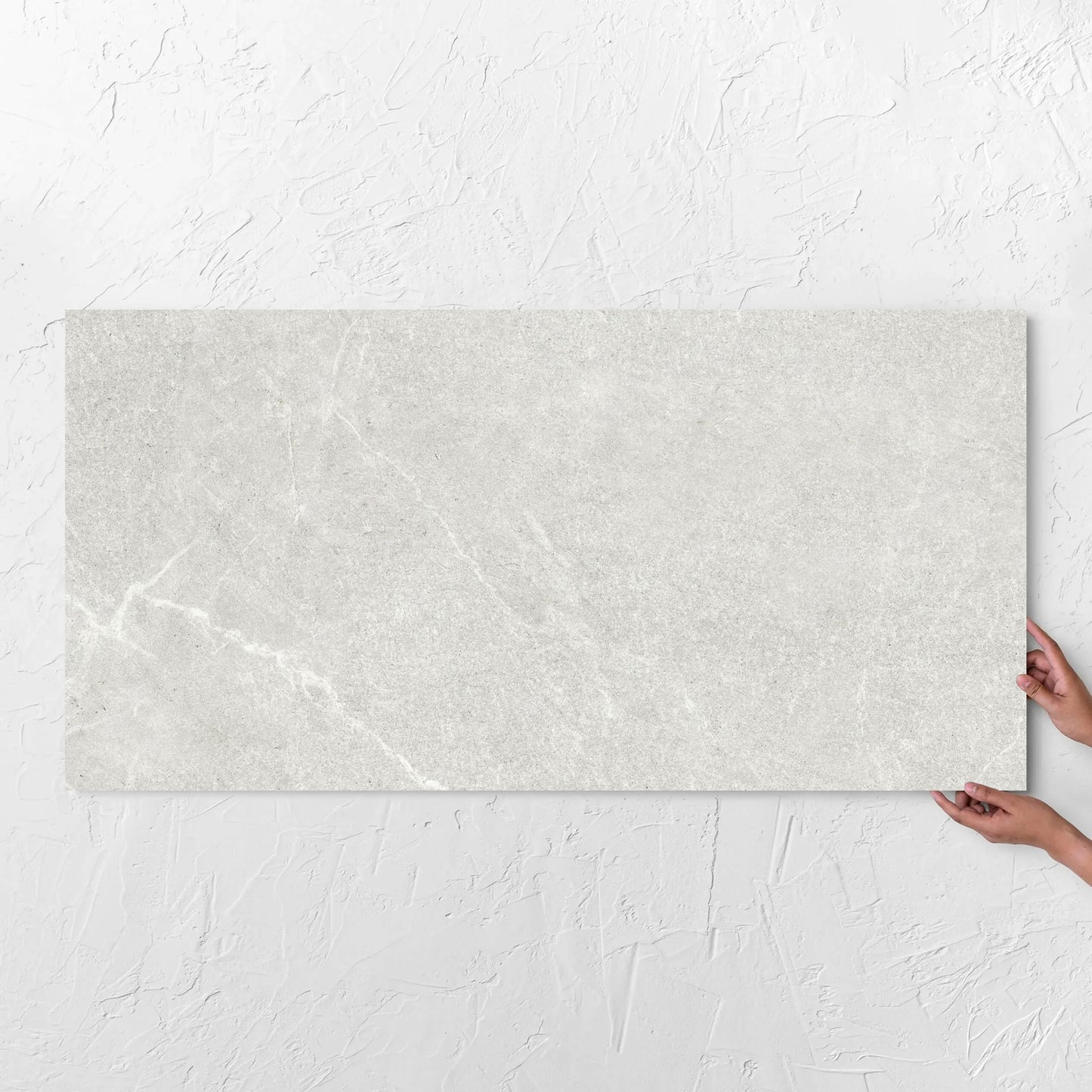 Hands holding a Lava Warm Grey Amber 600x1200mm tile against a white rendered wall, showing the warm grey surface