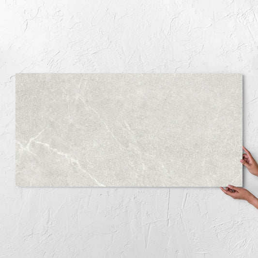 Hands holding a Lava Warm Grey Amber 600x1200mm tile against a white rendered wall, showing the warm grey surface