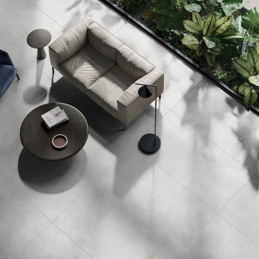 Lava White Amber 300x600mm porcelain floor tiles installed in a modern living room with a cream sofa, dark round coffee table, and floor lamp beside a glass wall with lush greenery.