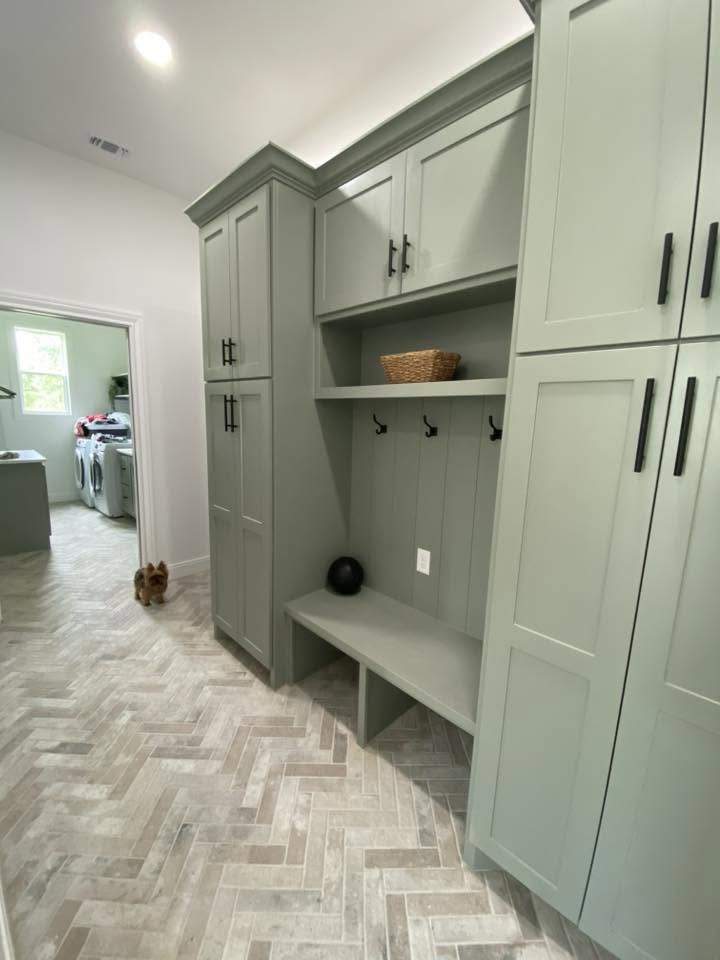London Fog Brick Tile 60x250mm laid in a herringbone pattern on a mudroom floor featuring sage green built-in cabinetry, bench seating, and an adjacent laundry room.