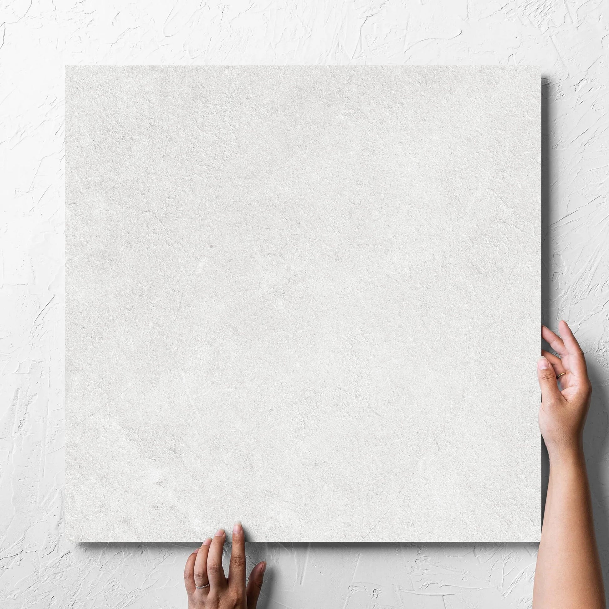 Hand holding a single Lusso Bianco Amber 600x600mm tile against a white rendered wall, showing the matt surface
