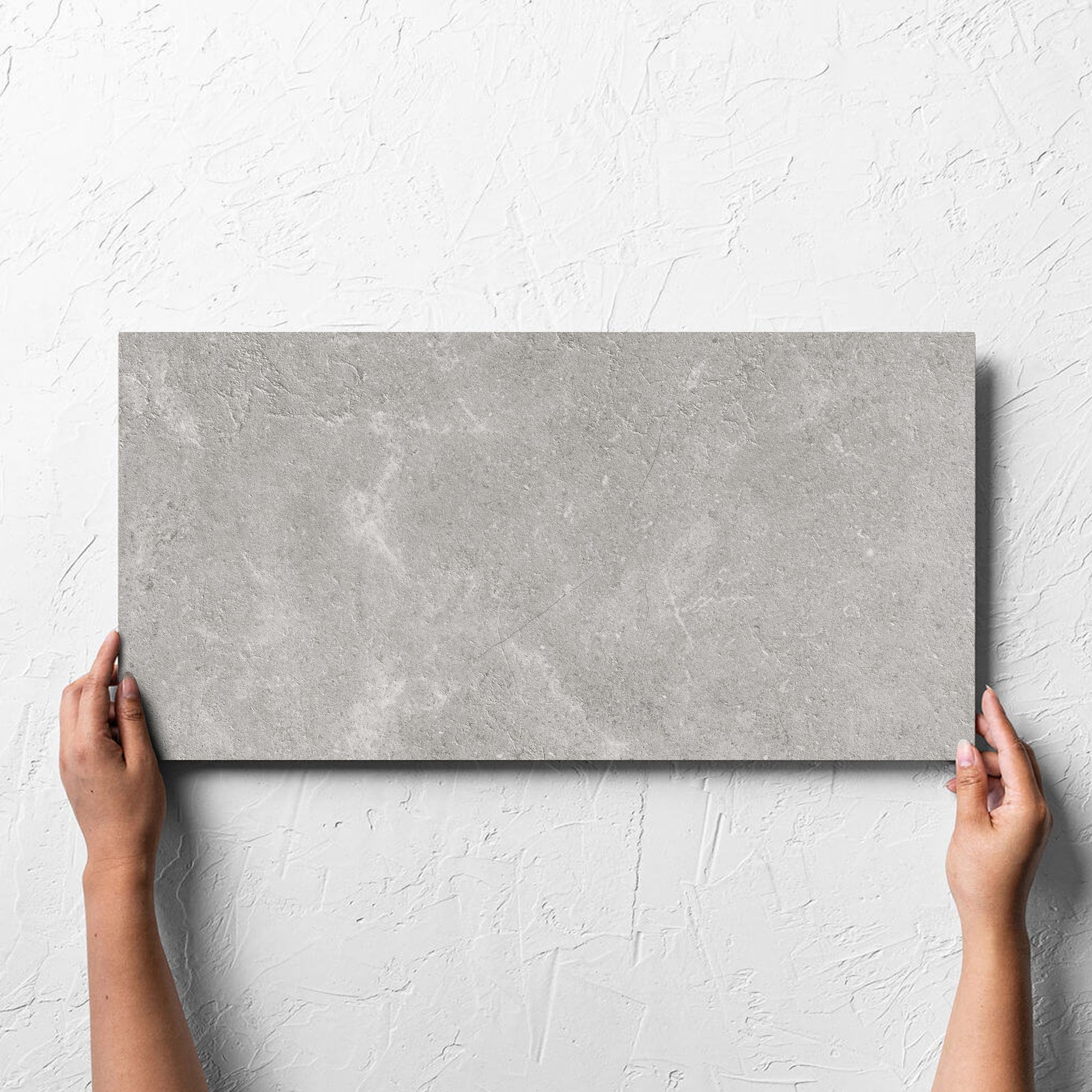 Hand holding a single Lusso Grigio Amber 300x600mm tile against a white rendered wall, showing the matt surface