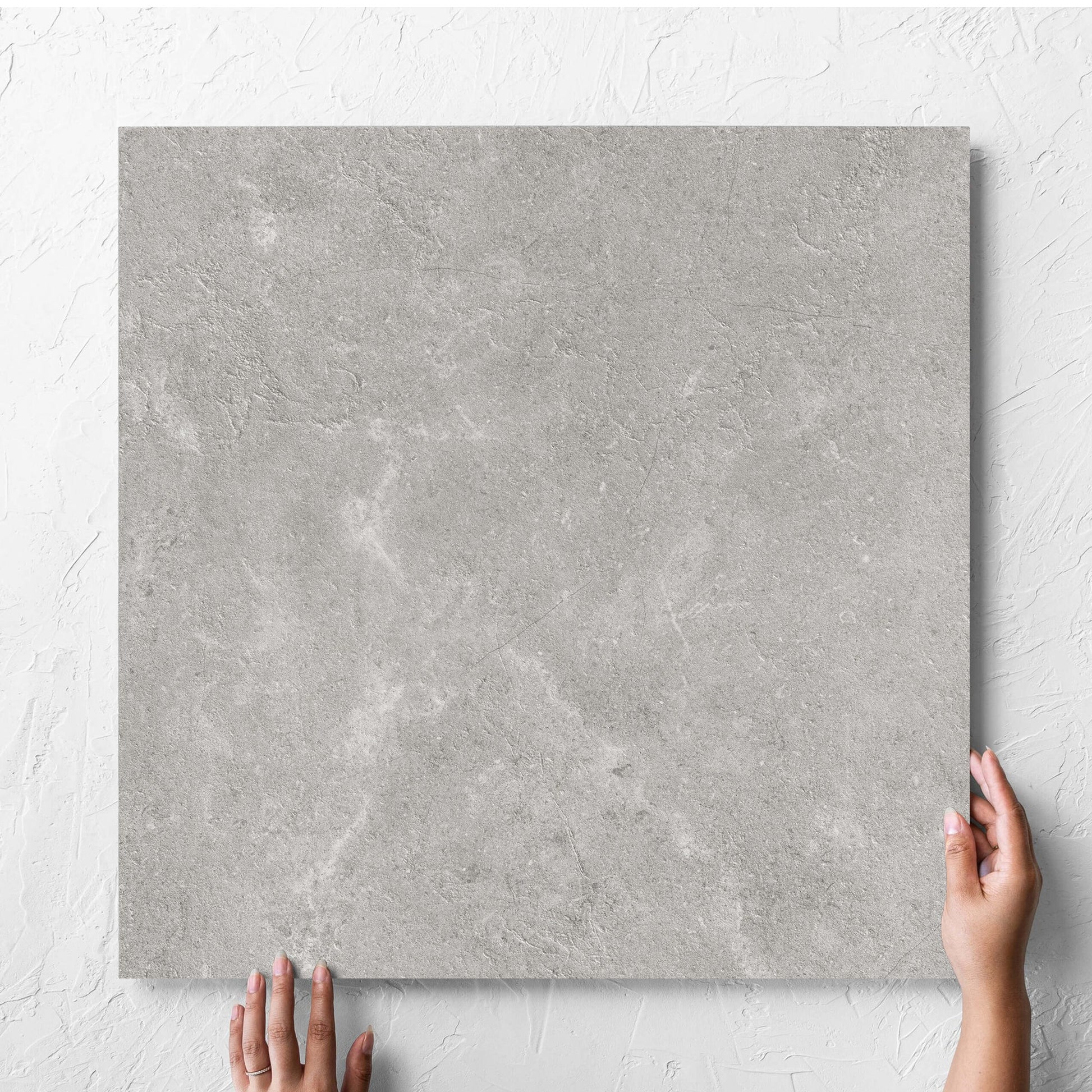 Hand holding a single Lusso Grigio Amber 600x600mm tile against a white rendered wall, showing the matt surface