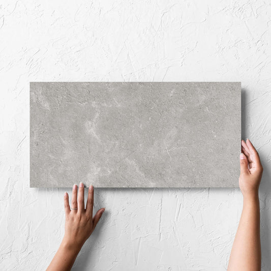 Hand holding a single Lusso Grigio Matt 300x600mm tile against a white rendered wall, showing the matt surface
