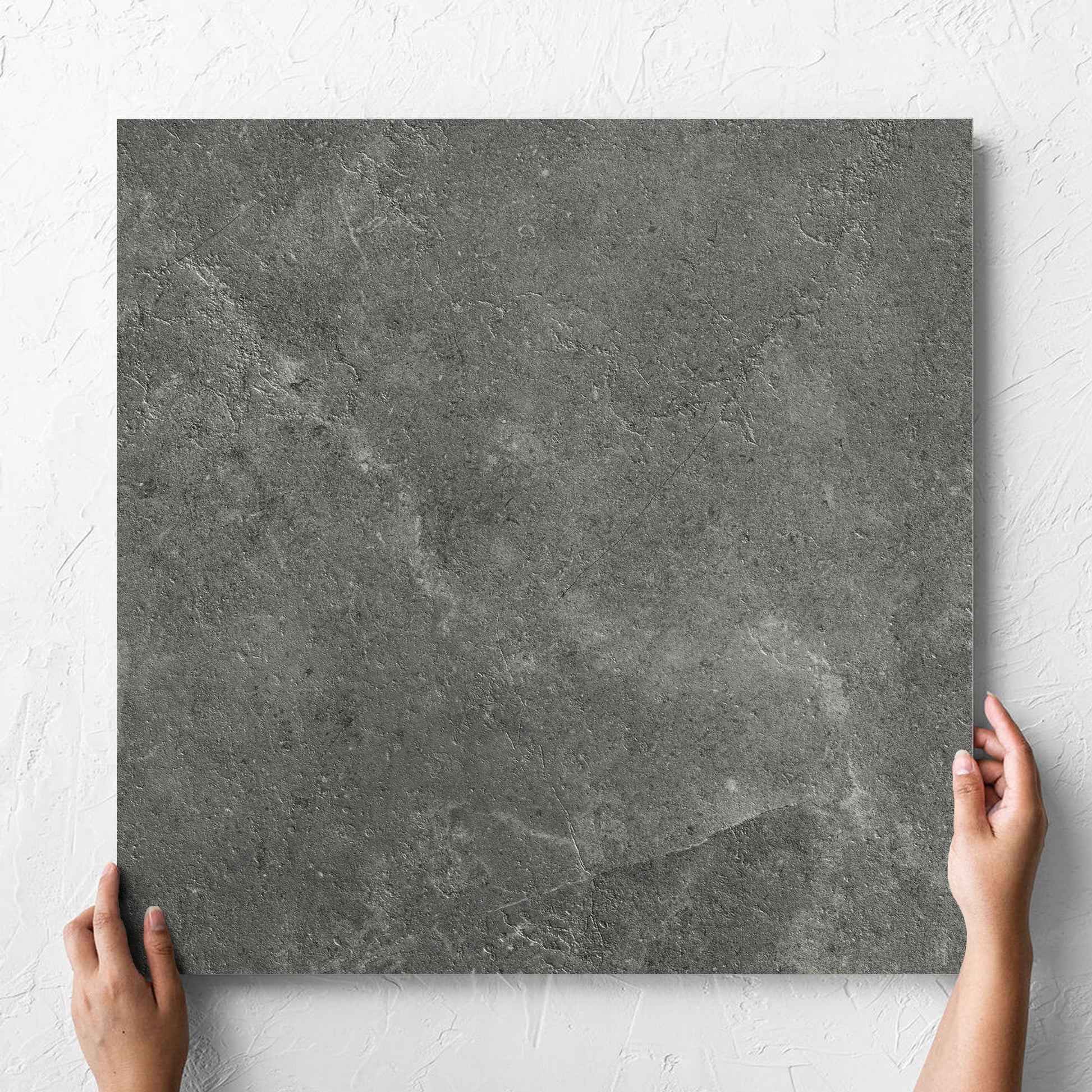 Hand holding a single Lusso Midnight Amber 600x600mm tile against a white rendered wall, showing the matt surface