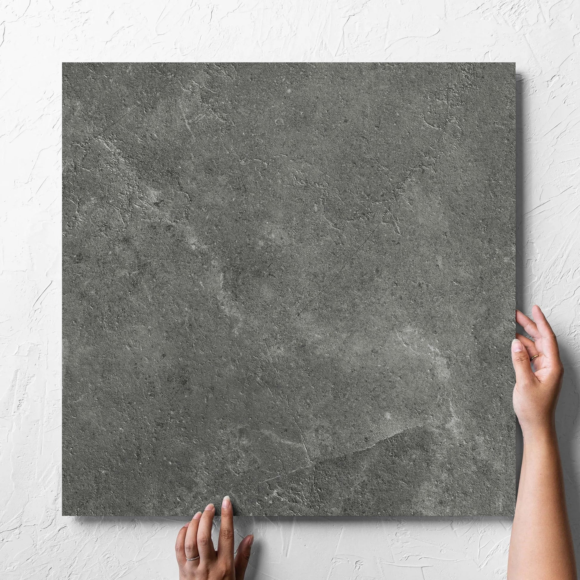 Hand holding a single Lusso Midnight Matt 600x600mm tile against a white rendered wall, showing the matt surface