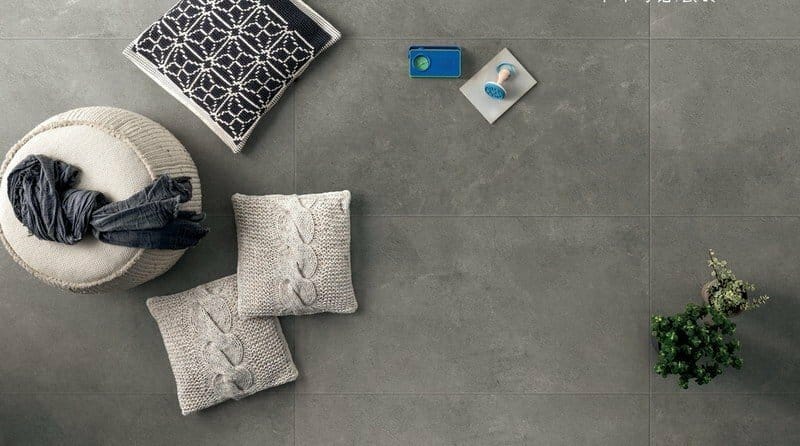 Lusso Nero Matt 600x600mm dark grey floor tiles styled in a living space with knitted cushions, a woven ottoman, and a small potted plant.