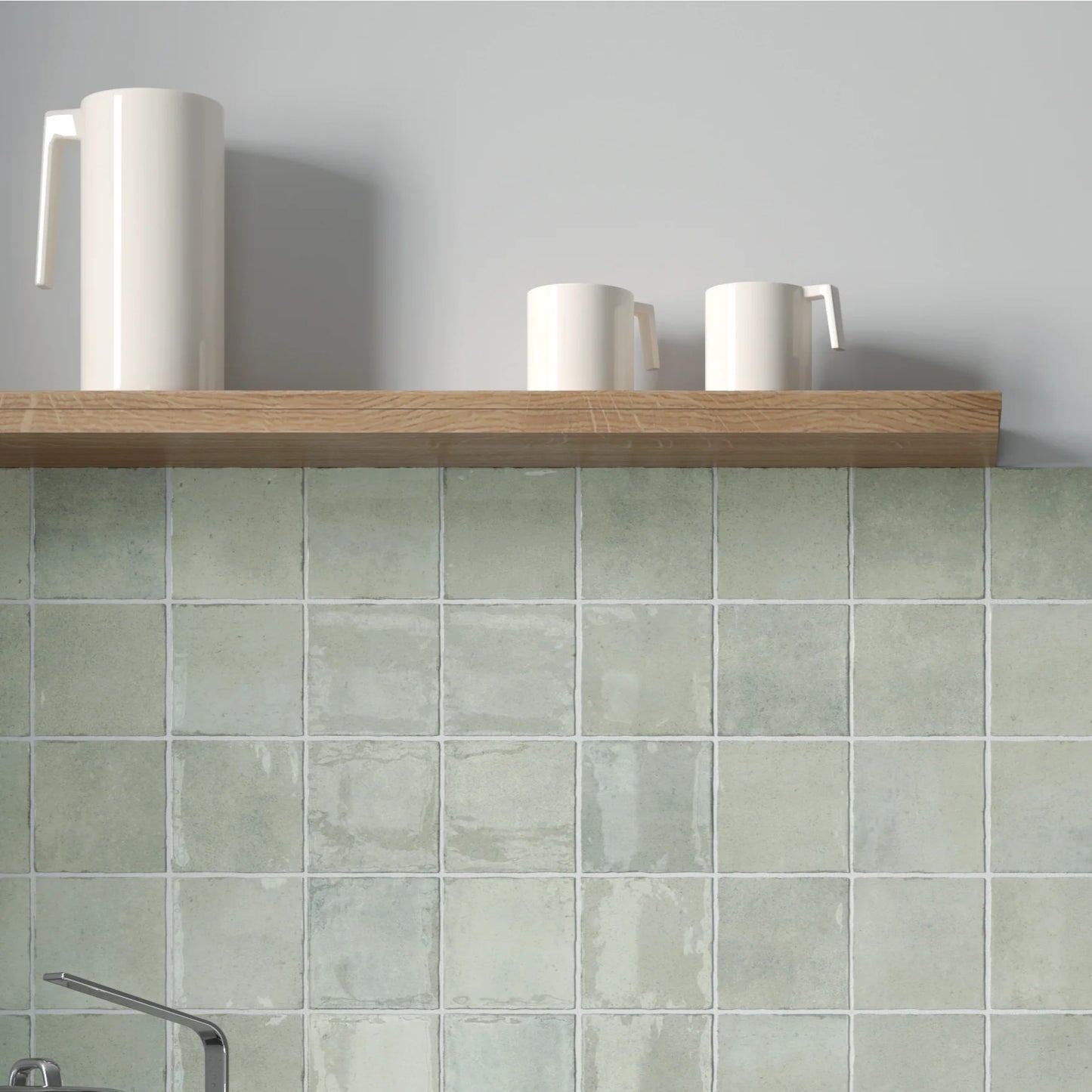 Majorca Gloss Cement 100x100mm sage green wall tiles installed as a kitchen splashback beneath a wooden shelf styled with white ceramic mugs and pitcher.