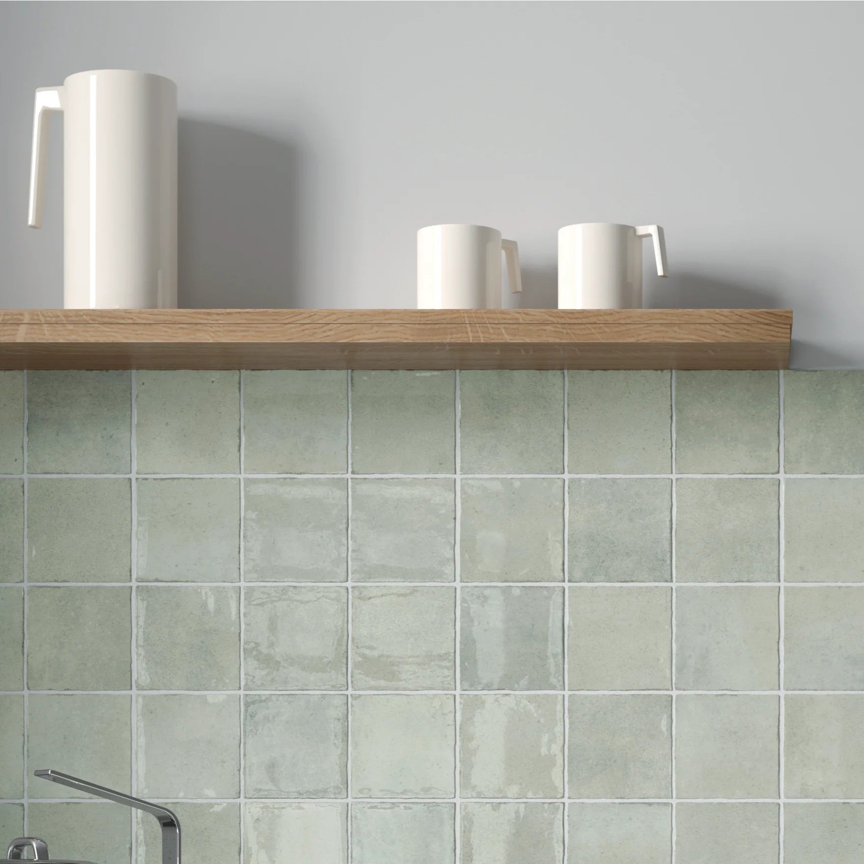 Majorca Gloss Cement 100x100mm sage green wall tiles installed as a kitchen splashback beneath a wooden shelf styled with white ceramic mugs and pitcher.