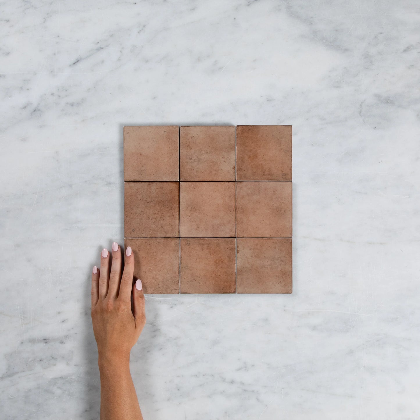 Nine Majorca Matt Cotto 100x100mm terracotta-toned square tiles arranged in a 3x3 grid on marble, with a hand shown for scale.
