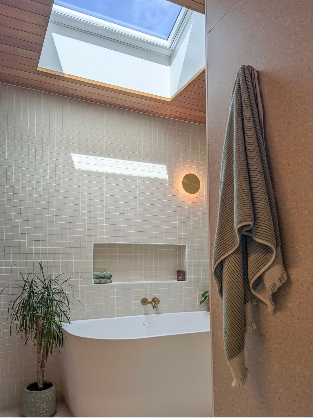 Marlowe Bone Matt Mosaic 48x48mm tiles covering a bathroom feature wall behind a freestanding bathtub with brass fittings, skylight, and timber ceiling.