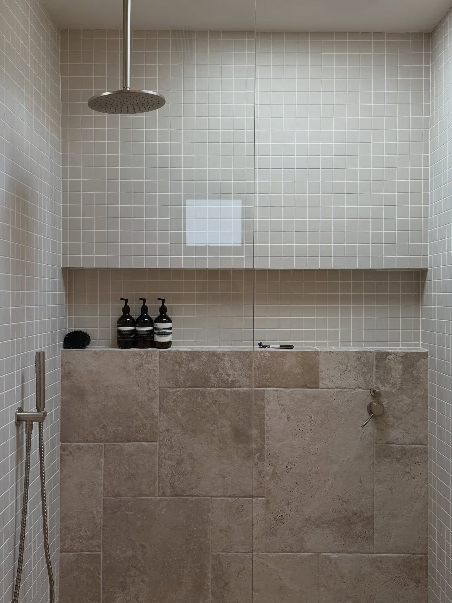 Marlowe Bone Matt Mosaic 48x48mm tiles covering shower walls above large-format travertine lower panels, styled with a ceiling-mounted rain head and Aesop products on a recessed niche shelf.