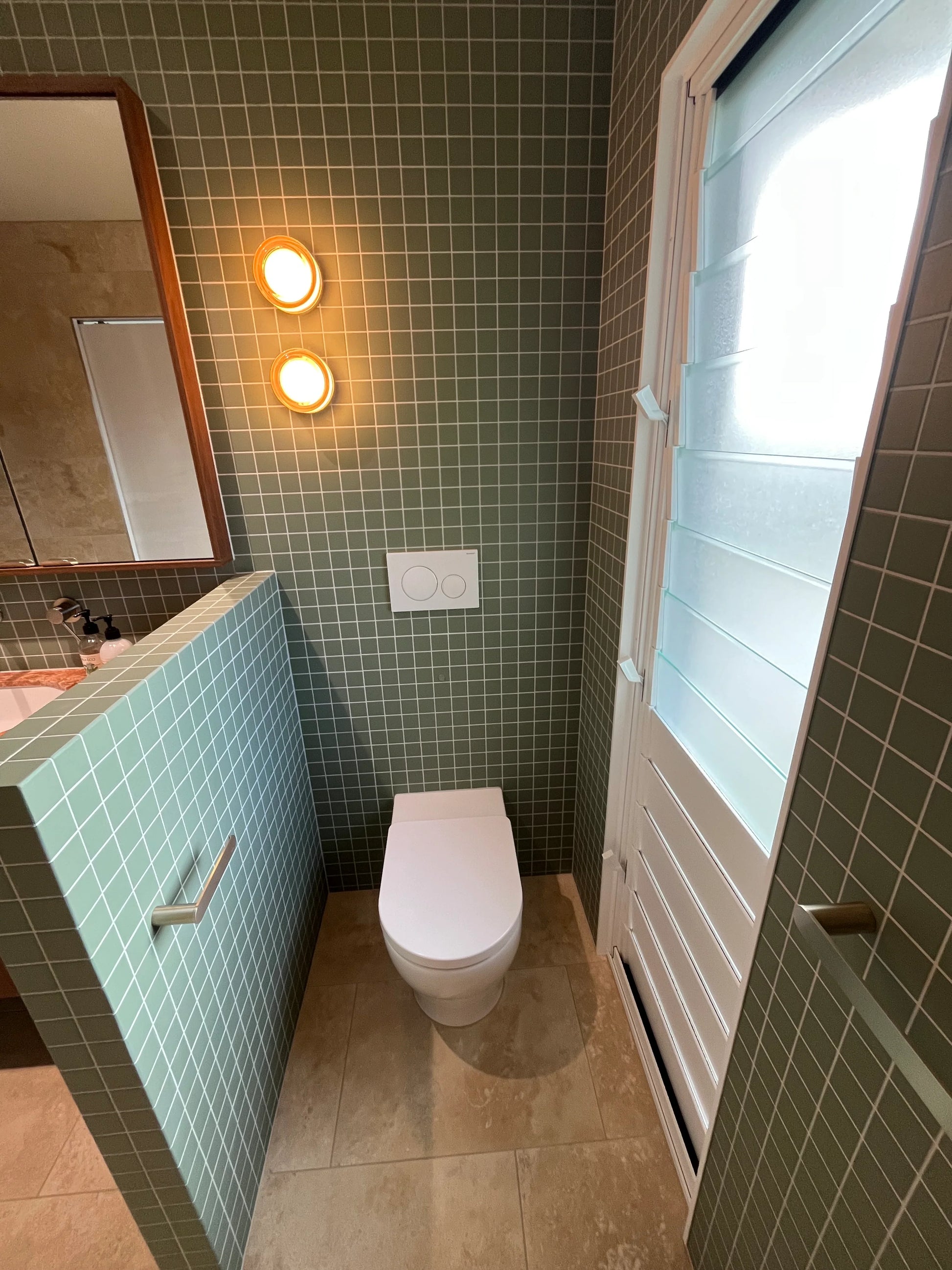 Marlowe Pistachio Green Matt Mosiac 48x48mm tiles covering a bathroom wall behind a wall-hung toilet, styled with brass wall lights, a timber-framed mirror, and frosted glass door.