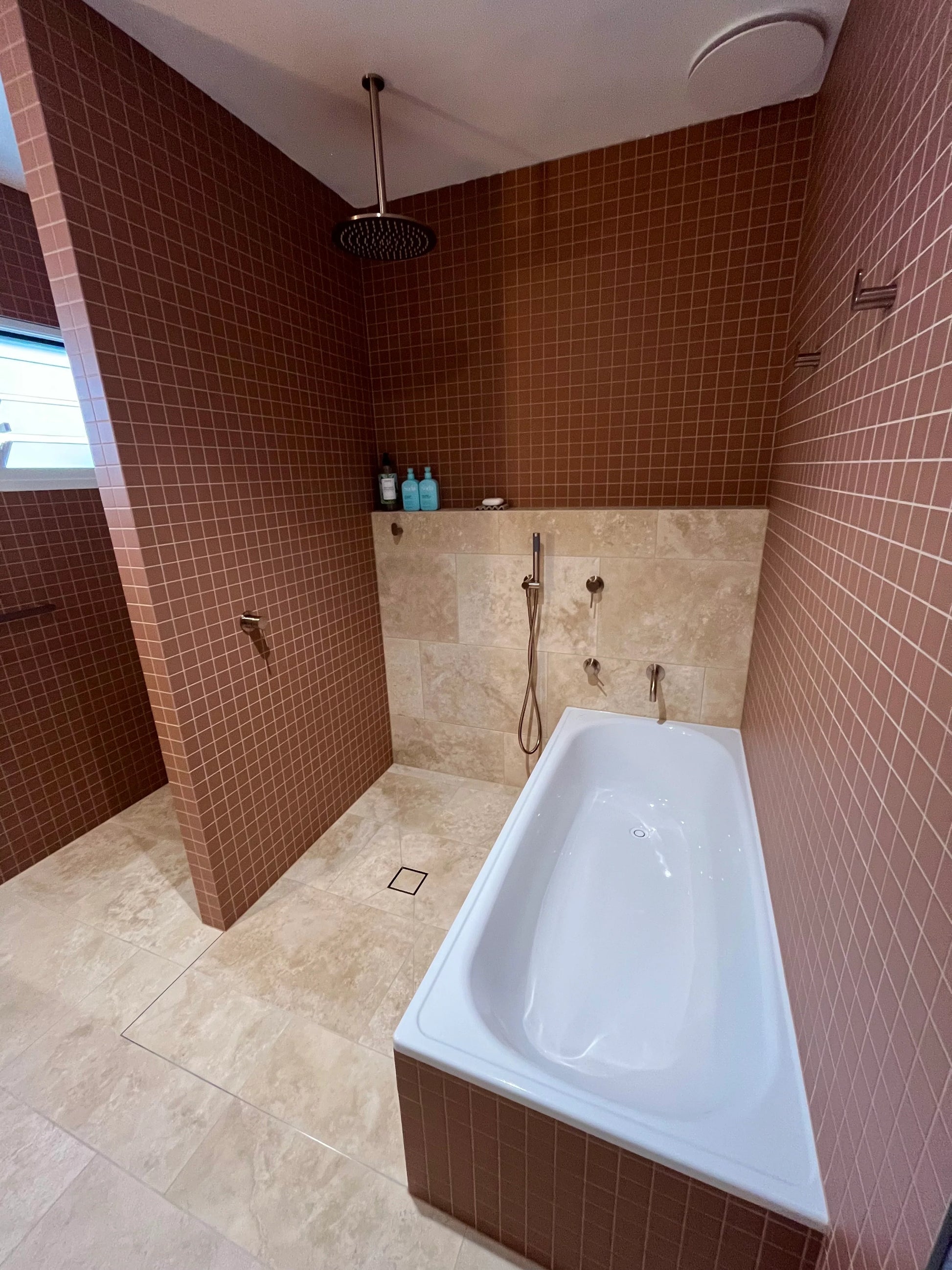 Marlowe Terracotta Matt Mosaic 48x48mm tiles covering bathroom walls alongside a freestanding white bath, ceiling-mounted rain shower head, and travertine floor tiles.