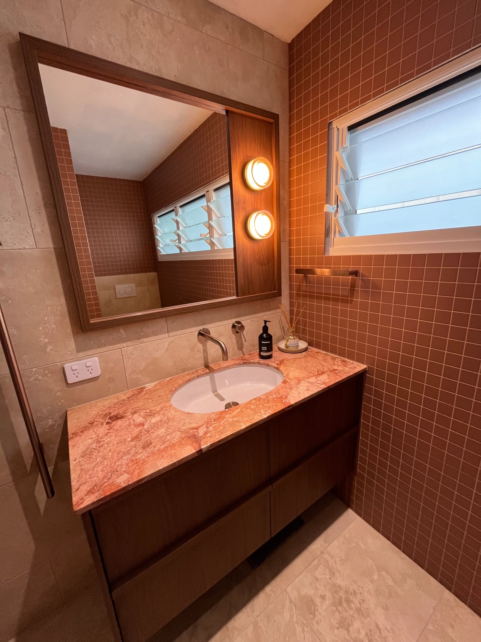 Marlowe Terracotta Matt Mosaic 48x48mm tiles covering a bathroom feature wall beside a pink marble vanity with timber cabinetry and round wall sconces.