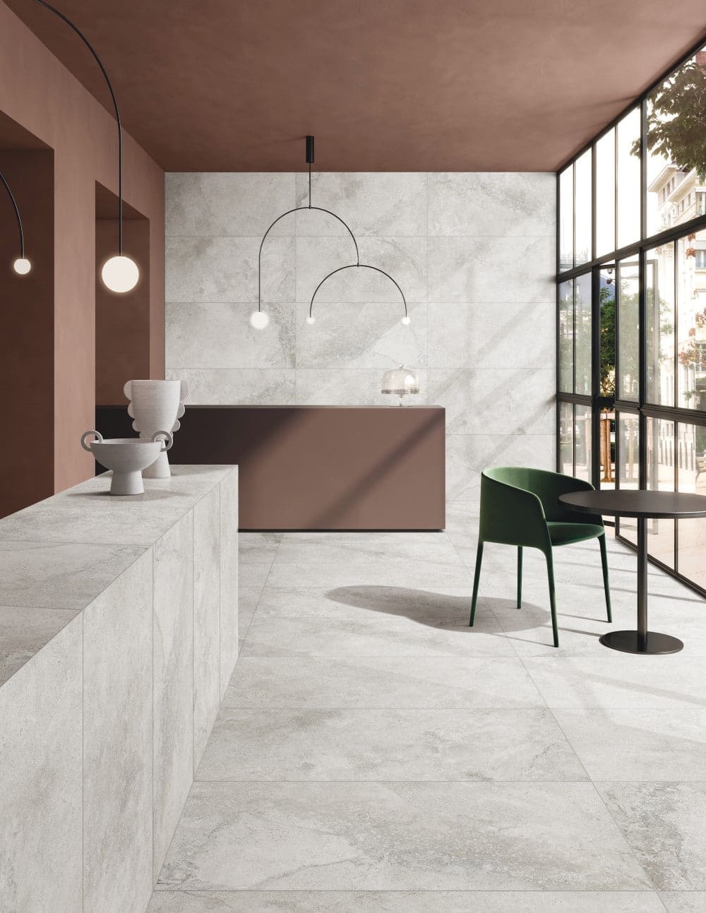 Mars Light External Pencil Bullnose 390.80x600x20mm light grey stone-look tiles covering the floor and feature wall of a contemporary lobby reception with terracotta ceiling and green armchair.