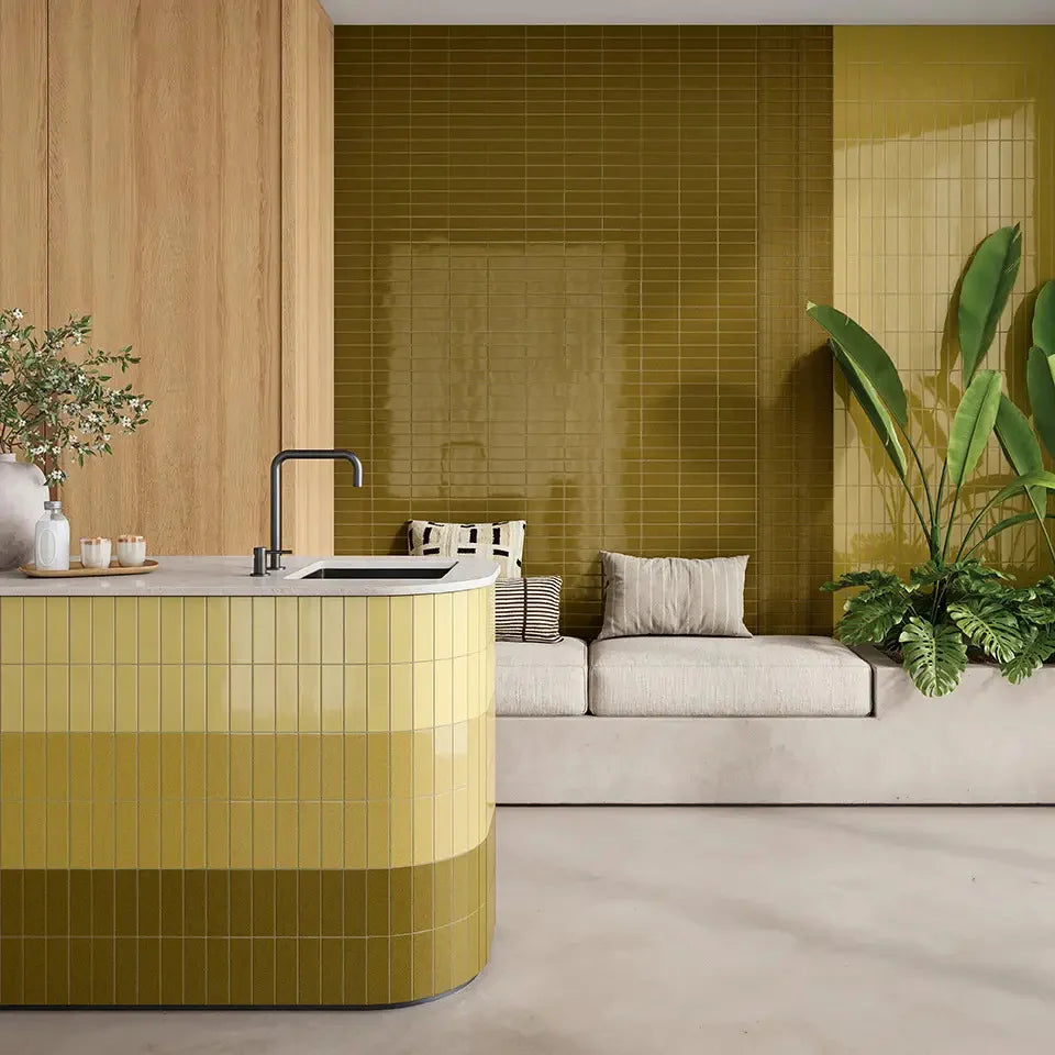 Micare Canale Alpi Dark Gloss 50x150mm olive-green tiles covering a curved kitchen island and feature wall beside a neutral sofa and tropical plants.