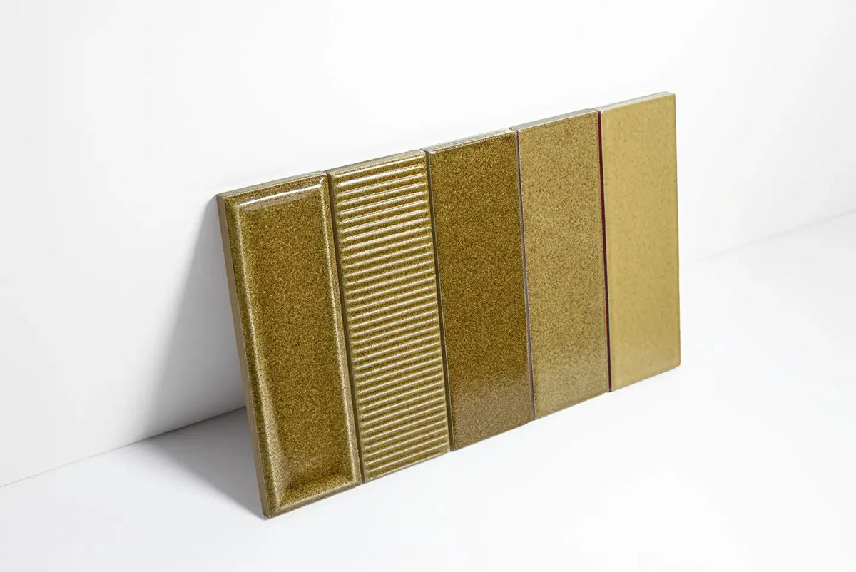Four Micare Canale Alpi Dark Gloss 50x150mm ceramic tiles leaning against a white wall, showcasing bevelled, ribbed, smooth, and flat gloss finishes in an olive gold tone.