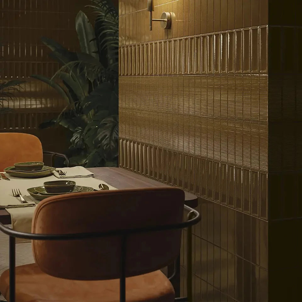 Micare Canale Ambar Dark Gloss 50x150mm tiles clad a warm amber restaurant booth wall beside leather chairs, tropical plants, and a brass wall sconce.