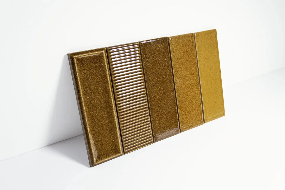 Micare Canale Ambar Dark Gloss 50x150mm tile sample board displaying four surface variations including bevelled, ribbed, speckled, and smooth finishes in warm amber and olive tones.
